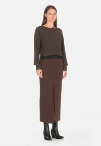 Maglione lavorato a maglia marrone con bordo in pizzo nero all'orlo, abbinato a una lunga gonna marrone con spacco frontale. La modella indossa stivali neri con tacco.