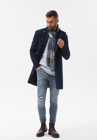 Mørkeblå uld overfrakke med store knapper, parret med en lyseblå sweater og et gråt ternet tørklæde. Båret med lyse slidte jeans og brune støvler.