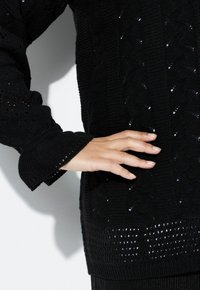 Černý pletený svetr s texturovaným vzorem, žebrovanými manžetami a volnými detaily podél lemu a rukávů.