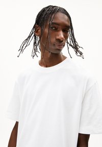 Weißes Baumwoll-T-Shirt mit kurzen Ärmeln und rundem Halsausschnitt. Der Stoff ist glatt und weist keine Muster oder Grafiken auf. Das Model hat geflochtenes Haar.