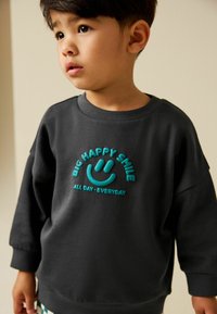 Tmavě šedá mikina s texturovanou tyrkysovou grafikou a nápisem „BIG HAPPY SMILE ALL DAY-EVERYDAY“ vytištěným pod ní.