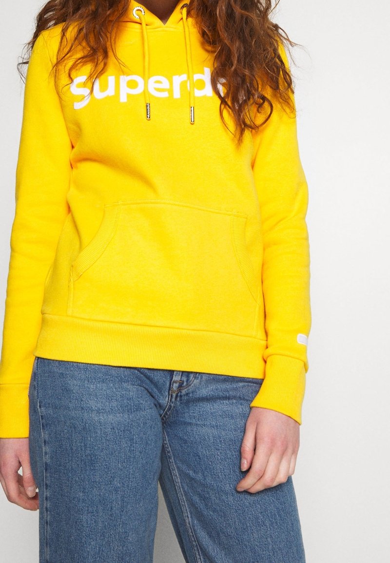 Femme portant un sweat à capuche jaune vif avec le texte blanc "Superd" et un jean bleu, debout devant un fond clair uni.