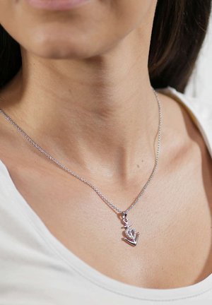 Silberne Halskette mit einem detaillierten Ankeranhänger, der eine strukturierte Oberfläche und dezenten Glanz aufweist, der auf nackter Haut über einem hellen Oberteil ruht.