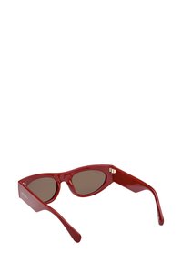 Röda cat-eye solglasögon med mörka linser och breda bågar, sedda från en låg vinkel mot en vit bakgrund.