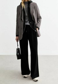 Blazer à carreaux noir et blanc sur un sweat à capuche noir, t-shirt gris, pantalon large noir et baskets blanches, tenant un sac noir.