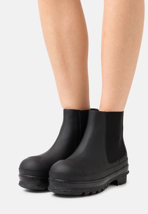 Black rubber ankle boots feature elastic side panels, rounded toe, textured sole, and a smooth surface with stitching details.