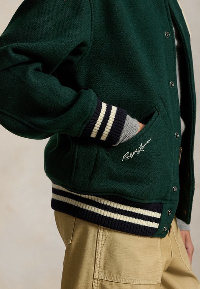 Zielona kurtka varsity z mieszanki wełny, z granatowymi i białymi paskami na ściągaczach, zatrzaskami oraz wyszywanym logo na kieszeni.