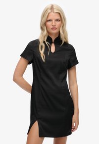 Schwarzes Kleid mit hohem Kragen, kurzen Ärmeln und einem Schlitz am Ausschnitt. Verfügt über einen seitlichen Schlitz und einen glatten, strukturierten Stoff.