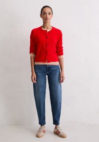 Gilet en maille rouge avec fermeture à boutons, poignets rayés. Associé à un jean en denim bleu avec une coupe décontractée et des baskets beiges avec des accents rouges.