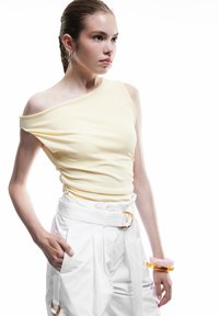 Top jaune clair à une épaule avec un tissu texturé, associé à un pantalon blanc taille haute avec une large ceinture et une grande boucle. Les accessoires comprennent des bracelets empilés en rose et orange.