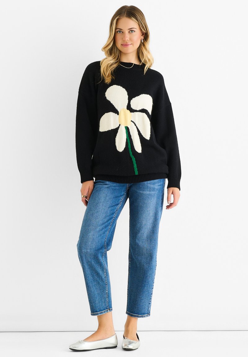 Maglione nero lavorato a maglia con un grande motivo floreale bianco, abbinato a jeans azzurri chiari e ballerine argento. Vestibilità rilassata, polsini a coste.