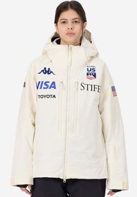 Giacca da sci color crema con cappuccio, caratterizzata da loghi blu e neri, tasche con zip e una vestibilità comoda. Realizzata in un materiale impermeabile.
