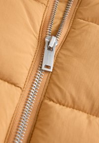 Gros plan d'une fermeture éclair en métal argenté sur un tissu matelassé beige, avec la fermeture éclair partiellement fermée et les coutures visibles.