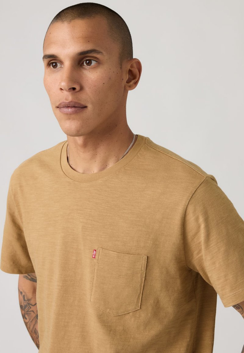 Young man with short hair wearing a light brown T-shirt with a chest pocket and visible tattoo on his left arm, neutral background.