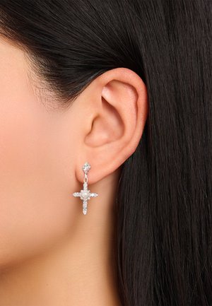 Nahaufnahme eines Ohrs einer Person mit glattem, dunklem Haar, die einen silbernen, kreuzförmigen Ohrring trägt, der mit kleinen Diamanten und einer zentralen Perle verziert ist.