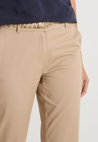 Pantalons beige avec une coupe droite, en tissu lisse. Comprends des poches avant et une ceinture en or tressée à la taille.
