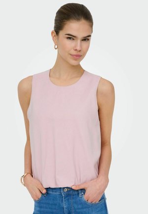 Sötét hajú nő rózsaszín ujjatlan felsőben és kék farmerruhában, kezét zsebében tartja, arany karika fülbevalót és karkötőt visel, világos háttér.