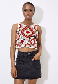Top court en crochet crème avec des motifs géométriques rouges, oranges et bruns ; associé à une jupe en denim noire avec des poches avant.
