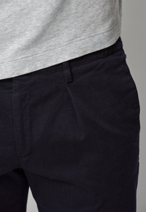 Pantalones azul marino oscuros hechos de una tela resistente, con pliegues frontales y un acabado suave, combinados con una camisa gris claro a la altura de la cintura.