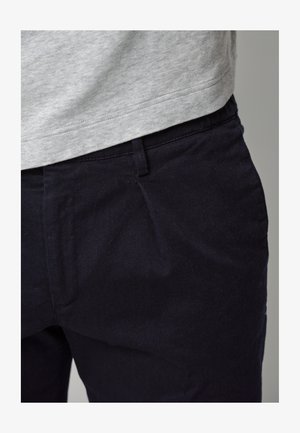 Dunkelblaue Hose aus robustem Stoff mit vorderen Falten und glatter Oberfläche, kombiniert mit einem hellgrauen Hemd auf Taillenhöhe.