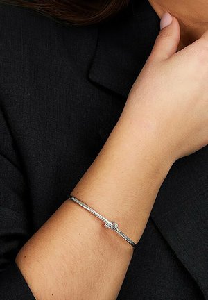 Silberner geflochtener Armreif mit Knotendetail. Der Armreif hat eine glatte, metallische Oberfläche und wird an einem Handgelenk vor einem dunklen Stoffhintergrund getragen.