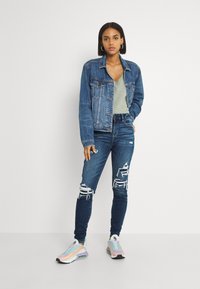 Casaco de ganga com um design clássico, na cor azul claro, e bolsos. Usado sobre um top verde e combinado com jeans skinny desgastados e ténis coloridos.