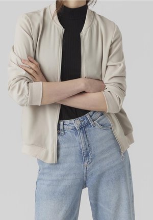 Persoon die een beige jas met rits draagt over een zwart topje met gekruiste armen, gecombineerd met lichtblauwe hoge taille jeans tegen een effen achtergrond.