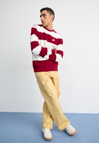 Sudadera a rayas rojas y blancas con el logo de Nike, combinada con pantalones amarillos de pierna ancha y zapatillas blancas con suela de goma, sobre un fondo azul.