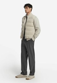 Beige gesteppte Puffer-Weste über einem grauen Langarmshirt, kombiniert mit dunkelgrauen Hosen und hellbraunen Sneakern. Glatte Texturen und neutrale Töne.