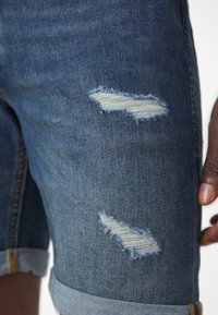 Jeansshorts in Mittelblau mit ausgefransten Rissen und umgeschlagenen Säumen. Verfügt über eine glatte Textur und sichtbare Nähte in der Nähe des Bundes.
