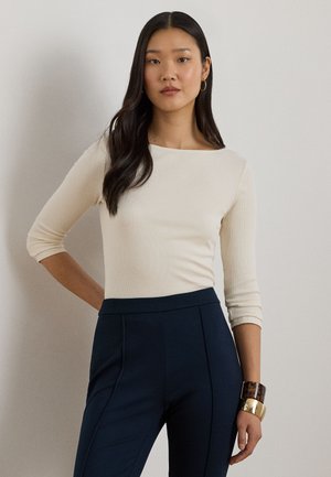 Kreminės spalvos ilgomis rankovėmis megztinis su valtimi kaklu, derinamas su tamsiai mėlynomis, aukštu liemeniu kelnėmis. Išskirtiniai auksiniai ir vėžlio kiauto spalvos apyrankės.