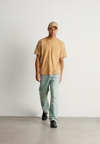 Μπεζ βαμβακερό μπλουζάκι με τσέπη, ανοιχτό μπλε τζιν και μαύρα παπούτσια. Το μοντέλο φοράει ένα ταιριαστό μπεζ καπέλο, όλα σε ουδέτερο φόντο.