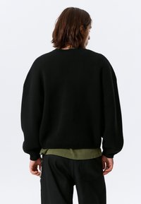 Pull en maille noire à coupe décontractée, épaules tombantes et ourlet côtelé. Superposé à une chemise vert olive, associé à un pantalon noir.