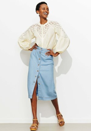 Femme souriante portant un chemisier en dentelle crème avec manches bouffantes, une jupe midi en denim bleu clair boutonnée et des sandales à brides marron clair.