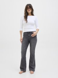 Maglietta bianca a coste con maniche lunghe abbinata a jeans flare grigio scuro a vita alta; la modella è in piedi a piedi nudi con lunghi capelli e occhiali da sole over-size.