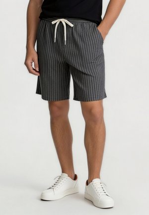 Mand iført sort og hvidt smaltstribet shorts med snøresnørre og hvide sneakers, stående med den ene hånd i lommen.