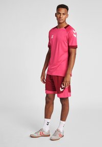 Rosa sporttröja med mörkröda accenter, i kombination med röda shorts. Har ett ventilerande tyg, logotyp på bröstet och signaturmönster på ärmarna.