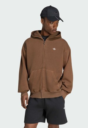Ung mand iført en brun hættetrøje med lynlås, sorte shorts og en sort Adidas kasket, kigger ned og mod sin højre side.