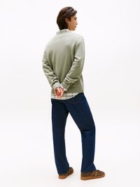 Zeleni pulover z rebrastimi manžetami, oblečen čez karo srajco, v kombinaciji s temnimi jeans hlačami in rjavimi čevlji. Model stoji z rokami za hrbtom.