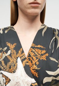 Femme portant un vêtement sombre à col en V avec un motif d'algues orange et beige ainsi que des étoiles de mer, visible du menton jusqu'au haut de la poitrine.