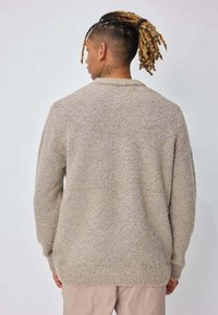 Personne aux cheveux nattés attachés en arrière portant un pull beige texturé et un pantalon clair, vue de dos sur un fond uni.