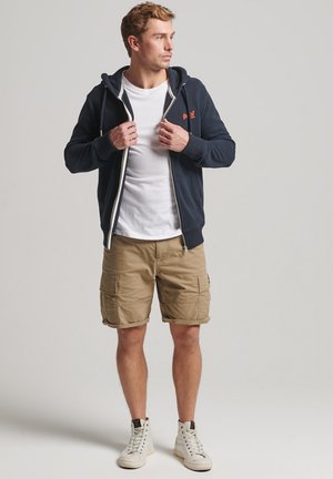 Felpa navy con zip e stringhe, t-shirt bianca e pantaloni cargo beige. Scarpe da ginnastica bianche completano il look. Un outfit semplice e casual.