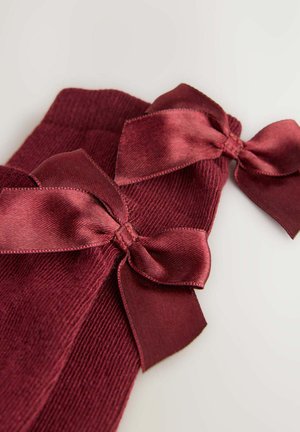 Une paire de chaussettes bordeaux à côtes avec de grands nœuds en satin sur les poignets, posées à plat sur un fond clair.