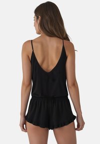 Jumpsuit sans manches noir avec un dos en V profond et une taille élastique, présentant des shorts à volants pour plus de texture et de détails. Finition en tissu lisse.