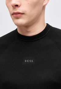 Εγγύς πλάνο ενός ατόμου που φορά μαύρο πουλόβερ με στρογγυλή λαιμόκοψη και τη λέξη "BOSS" τυπωμένη σε γκρι στο στήθος.