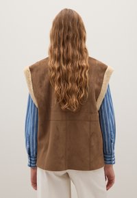 Braun ärmellose Weste mit einem weichen, Kunstfellkragen. Das Design umfasst eine Wildleder-Optik und einen geraden Schnitt, getragen über einem blau gestreiften Hemd.