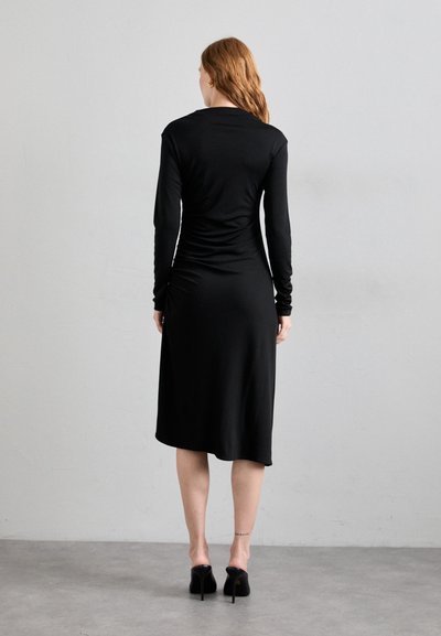 Une robe noire à manches longues avec un bustier ajusté, un drapé dans le dos et une longueur midi, fabriquée en tissu lisse et extensible, associée à des talons hauts.