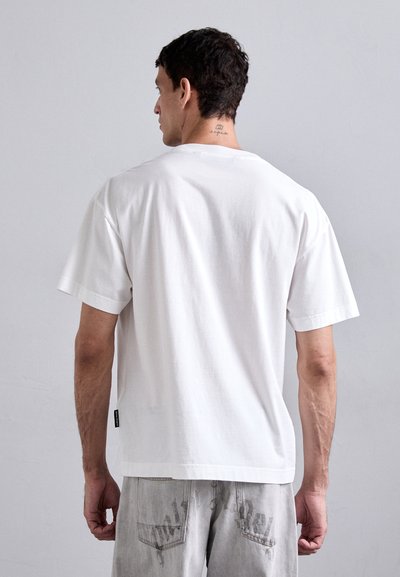 T-shirt en coton blanc à manches courtes, coupe décontractée, encolure ronde et petite étiquette de marque sur le côté. Design sobre, texture douce.
