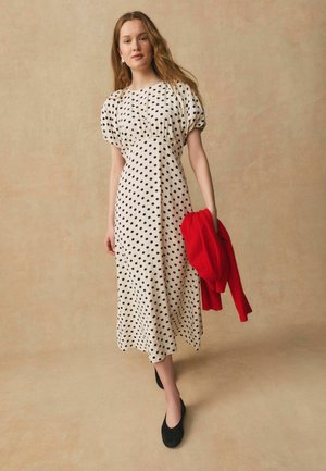 Femeie în rochie midi albă cu buline negre, ținând un pulover roșu, purtând pantofi negri, stând într-un decor bej.