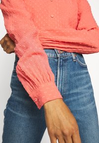 Donna che indossa una blusa a maniche lunghe in tessuto corallo con polsini abbottonati e jeans blu a vita alta, con la mano appoggiata vicino alla vita.